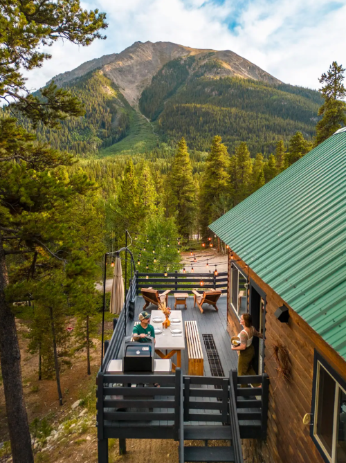 A scenic mountain view from a cabin deck with people dining and enjoying nature among tall trees.