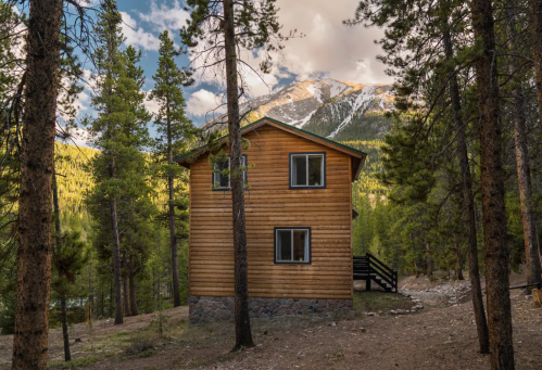 A wooden cabin nestled among tall trees, with mountains in the background under a partly cloudy sky.