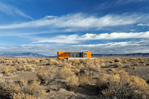 A modern house stands alone in a vast, arid landscape under a blue sky with scattered clouds.