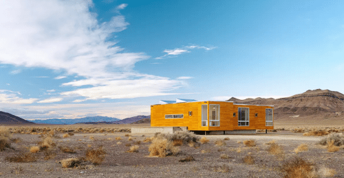 A modern wooden house sits on a barren landscape with mountains and a blue sky in the background.