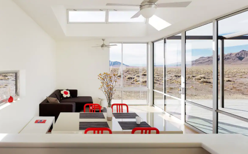Modern interior with a sofa, dining table, and large windows showcasing a desert landscape outside. Bright and airy atmosphere.