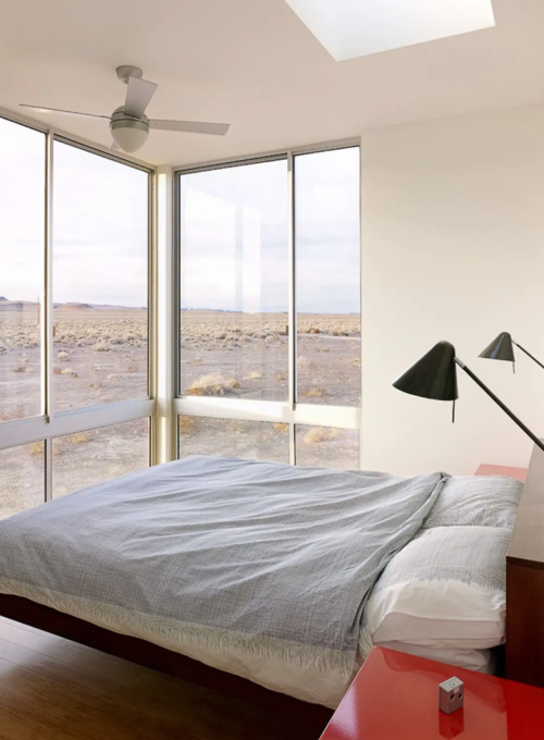 Modern bedroom with a large window overlooking a desert landscape, featuring a bed, ceiling fan, and stylish lamps.