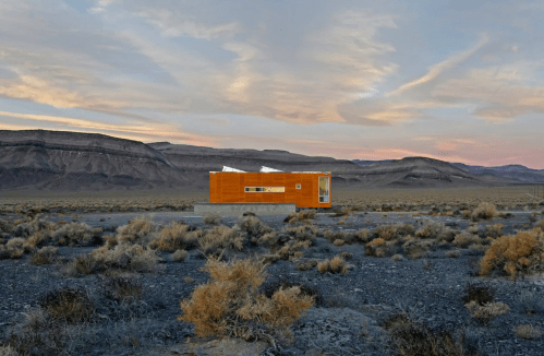 A modern house stands in a desert landscape, surrounded by sparse vegetation and mountains under a colorful sky.