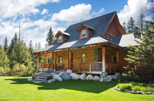 A rustic wooden cabin with a porch, surrounded by lush green grass and trees under a blue sky with fluffy clouds.
