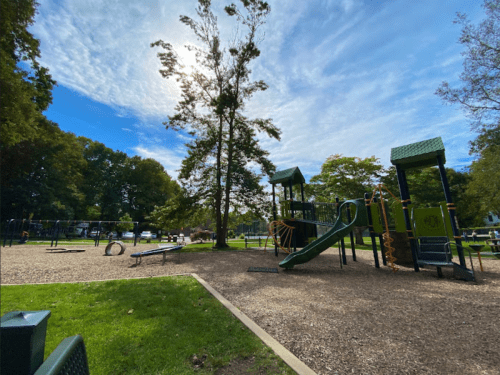 A playground with slides and climbing structures, surrounded by trees and a blue sky with scattered clouds.