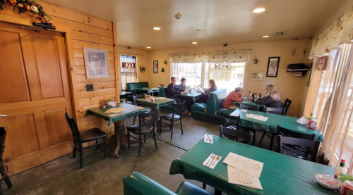 Cozy restaurant interior with green tables, patrons dining, and wooden decor. Bright natural light from windows.