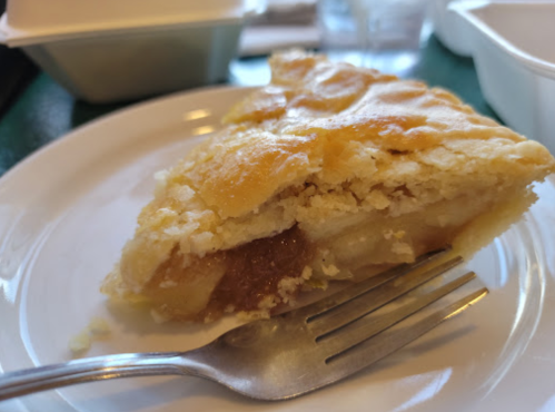 A slice of apple pie on a white plate with a fork beside it, showcasing a golden crust and apple filling.