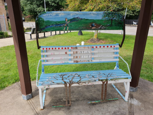 A colorful, artistic bench with a scenic mural on the backrest, surrounded by greenery in a park setting.