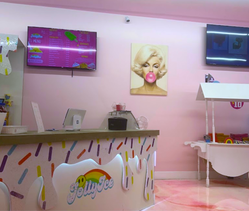 A colorful ice cream shop interior with a pink theme, featuring a menu board and a portrait of a woman blowing bubble gum.