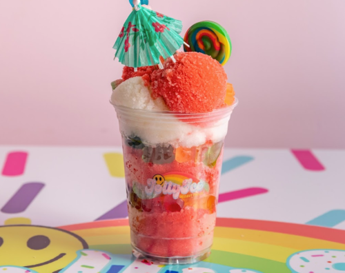 A colorful dessert cup filled with layers of ice, candy, and a small umbrella decoration against a pastel background.
