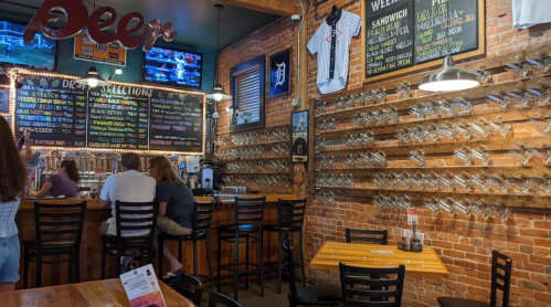 A cozy bar with wooden tables, a brick wall, and a menu board displaying drink selections. Patrons sit at the bar.