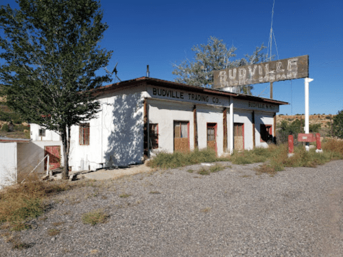 Abandoned Budville Trading Co. building with a weathered sign, surrounded by trees and gravel road under a clear blue sky.