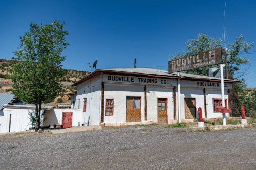 A weathered building with "Budville Trading Co." sign, surrounded by trees and a clear blue sky.