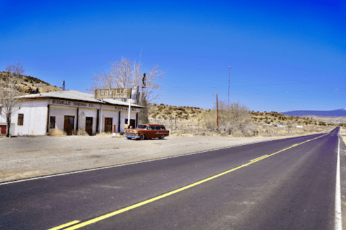 Abandoned building with a vintage car parked outside, alongside a deserted road in a rural landscape.