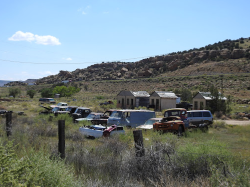 Abandoned vehicles and old buildings in a rural landscape with hills and blue sky.