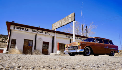 A vintage car parked in front of the Budville Trading Co. building in a desert setting. Clear blue sky above.