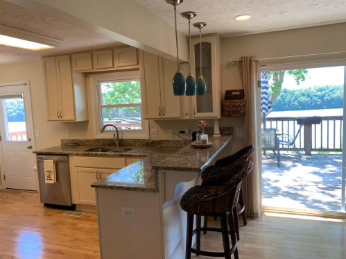 Bright kitchen with granite countertops, bar stools, and a view of a deck overlooking a lake. Natural light floods the space.