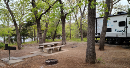 A campsite with a picnic table, fire pit, and an RV surrounded by trees near a lake.
