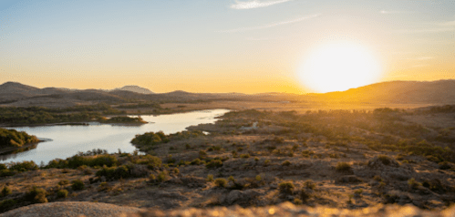 A serene landscape at sunset, featuring a river winding through hills and trees under a colorful sky.