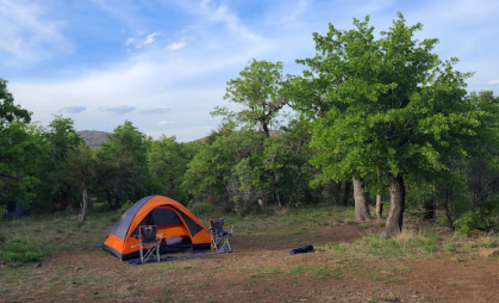 A camping scene featuring an orange and gray tent surrounded by green trees and two folding chairs.