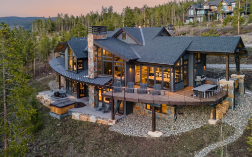 A modern mountain home with large windows, a wraparound deck, and surrounded by trees at sunset.