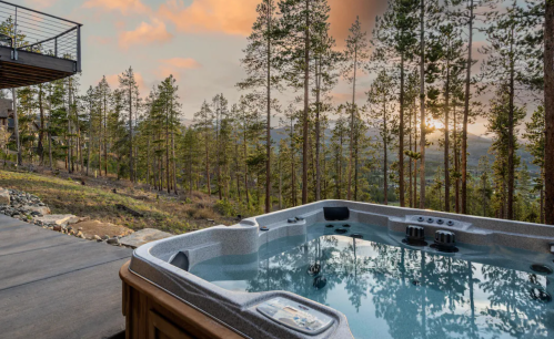 A hot tub on a patio overlooking a forested landscape at sunset, with trees and mountains in the background.
