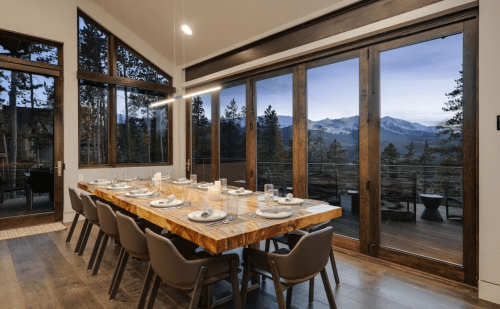 A modern dining room with a large wooden table set for dinner, featuring large windows with mountain views.