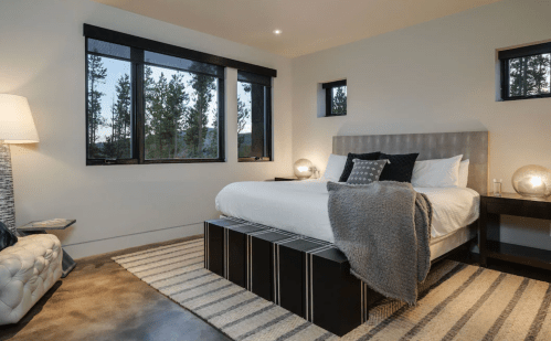 Modern bedroom with a gray bed, cozy blanket, and large windows showcasing trees outside. Soft lighting and a rug complete the look.