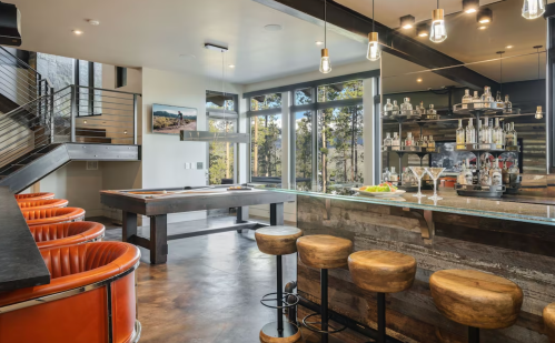 Modern bar area with wooden stools, orange chairs, a pool table, and large windows showcasing a forest view.
