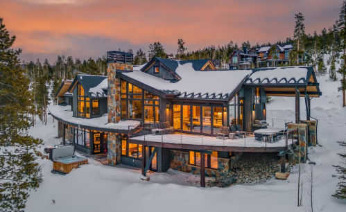 A modern mountain home with large windows, surrounded by snow and trees, under a colorful sunset sky.