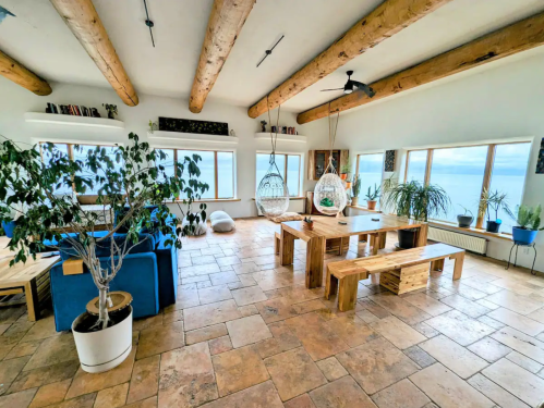Bright, airy living space with wooden beams, a large dining table, plants, and panoramic ocean views.