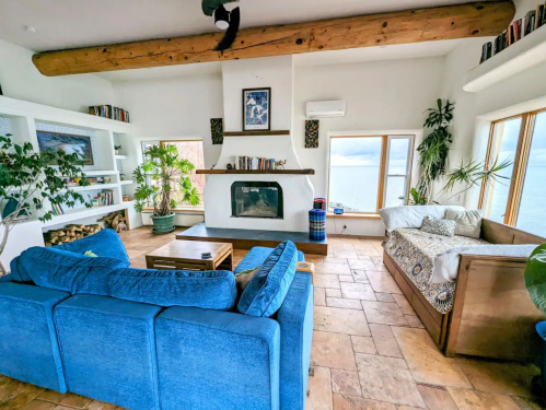 Cozy living room with blue sofas, a fireplace, large windows, and ocean views, featuring plants and wooden accents.