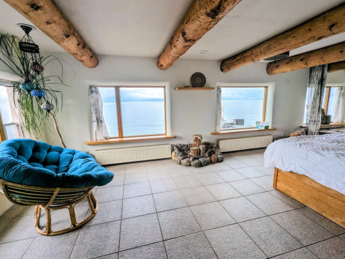 Cozy bedroom with large windows overlooking the sea, featuring a blue chair and wooden accents.
