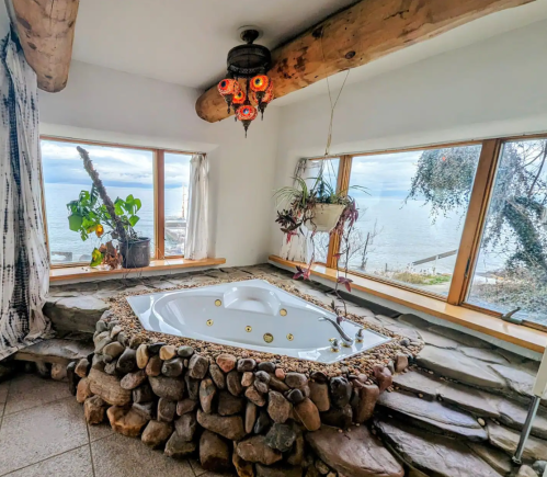 A cozy bathroom with a stone tub, large windows, and a view of the sea, featuring natural decor and warm lighting.