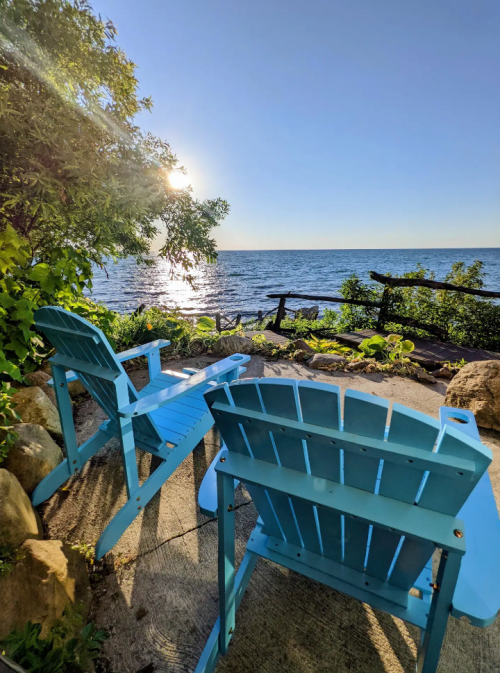 Two blue Adirondack chairs facing a serene lake under a clear sky, surrounded by greenery and sunlight.