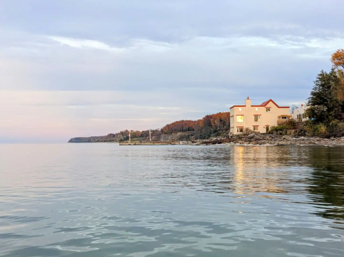 A calm lake scene at sunset, featuring a pink house along the shore and autumn-colored trees in the background.