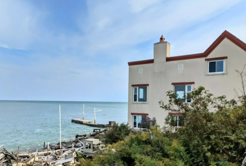 A coastal building beside a calm sea, with a wooden pier and scattered driftwood in the foreground.