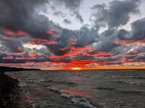 A vibrant sunset over a choppy lake, with dramatic clouds in shades of orange, red, and gray reflecting on the water.