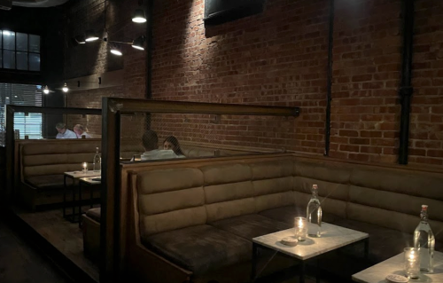 Cozy restaurant interior with brick walls, dim lighting, and private booths, featuring candles and water bottles on tables.