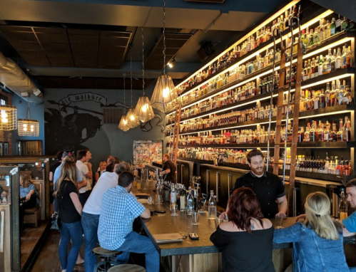 A lively bar scene with patrons at the counter, shelves filled with bottles, and warm lighting.