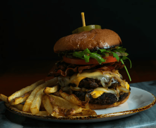 A towering burger with cheese, bacon, lettuce, and tomato, served with crispy fries on a plate.