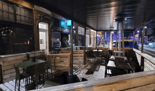 A cozy outdoor dining area with wooden seating and heaters, illuminated by street lights at night.