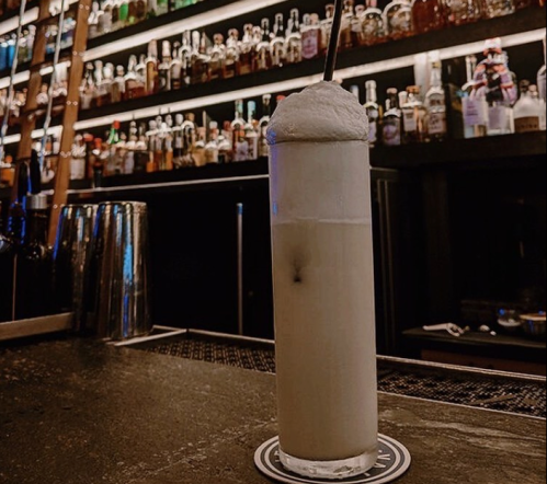A tall, frothy cocktail sits on a bar counter, with a well-stocked shelf of bottles in the background.
