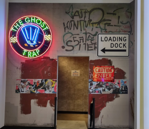 Neon sign reading "The Ghost Trap" above a loading dock entrance with graffiti and caution signs.