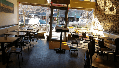 Brightly lit restaurant interior with wooden tables and chairs, featuring large windows and a view of the street outside.