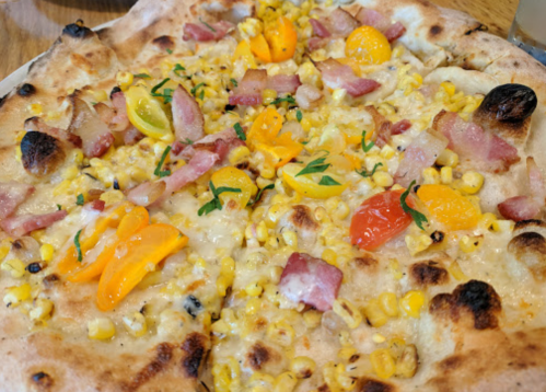 A close-up of a pizza topped with corn, bacon, cherry tomatoes, and herbs on a golden crust.