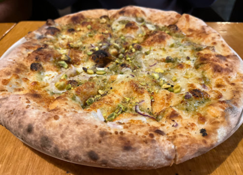 A close-up of a freshly baked pizza topped with cheese, pistachios, and herbs on a golden crust.