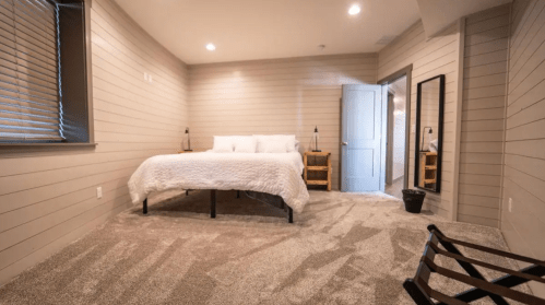 Cozy bedroom featuring a large bed, soft carpet, and natural light from windows, with a mirror and wooden furniture.