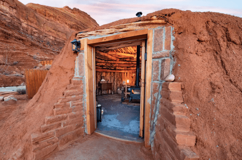 A rustic earthen structure with a wooden door, set against a rocky landscape at sunset. Inside, a warm glow is visible.