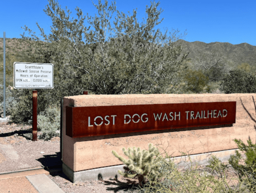 Sign for Lost Dog Wash Trailhead in a desert landscape, with mountains and vegetation in the background.